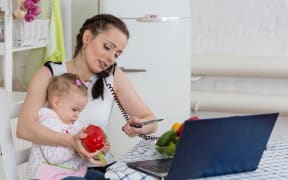 Photo of a multi-tasking mother who has a child on her knee, a computer in front of her and who is also trying to prepare vegetables for dinner