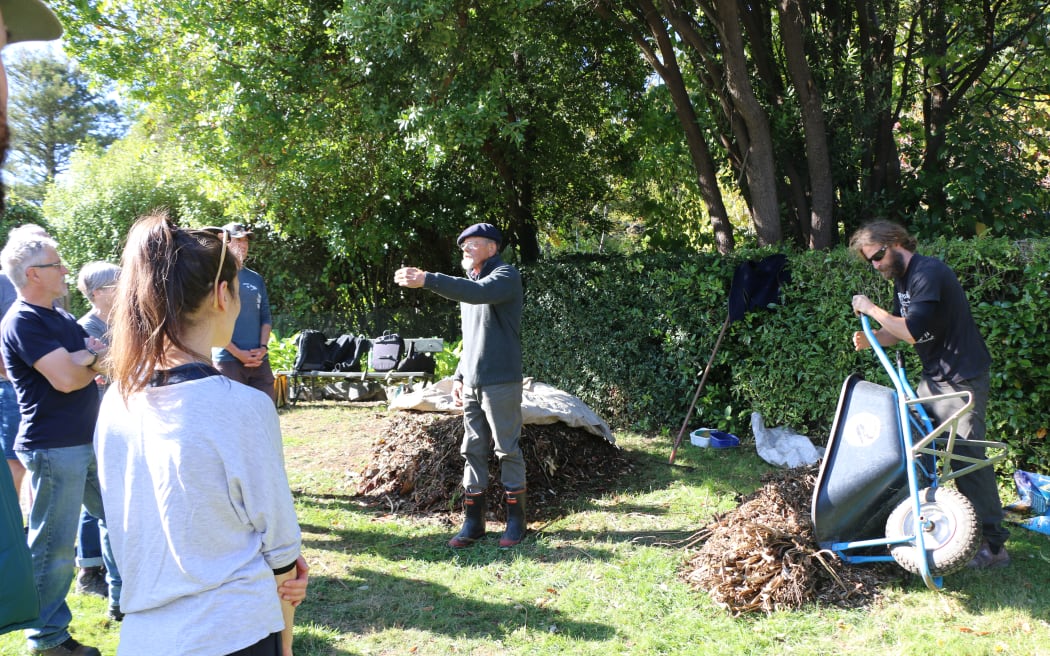 Jim is standing in front of a compost pile and talking to a small group of people. Another man is tipping a wheelbarrow full of compost onto the grass beside Jim.