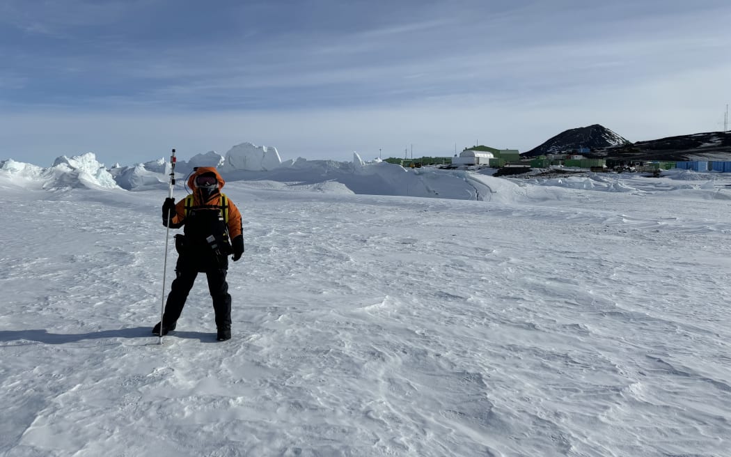 A person wearing black and orange cold-weather gear with the hood up stands on a flat expanse of ice criss-crossed with weaving patterns. In the background, jumbles of ice pile up. The buildings of Scott Base and black hills are visible in the background on the right, beneath a cloud-streaked blue sky.