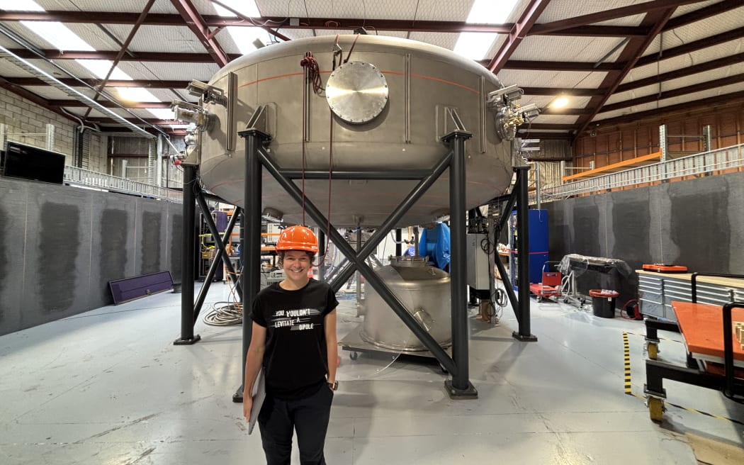 A person wearing an orange hard hat and a black t-shirt with the words 'You wouldn't levitate a dipole' - riffing off the 'You wouldn't steal a car' series of PSAs about movie pirating in the early 00s. Behind her, there is a large metal cylinder on a metal frame inside a warehouse.
