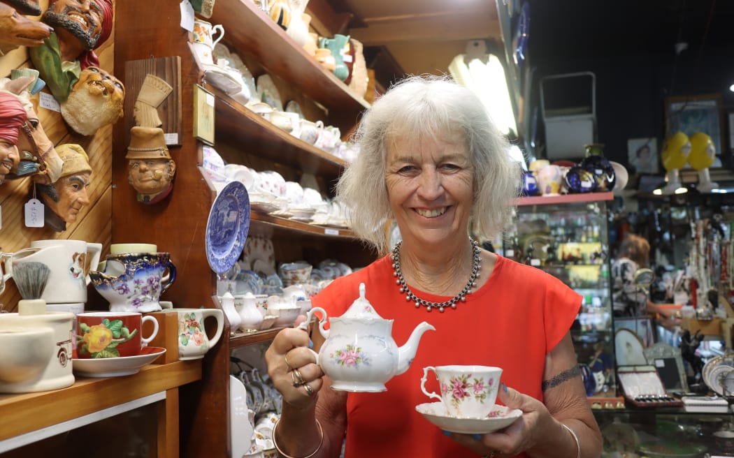 Picture of a woman with grey hair, standing in front of a wall of teacups, holding a teacup and tea pot.