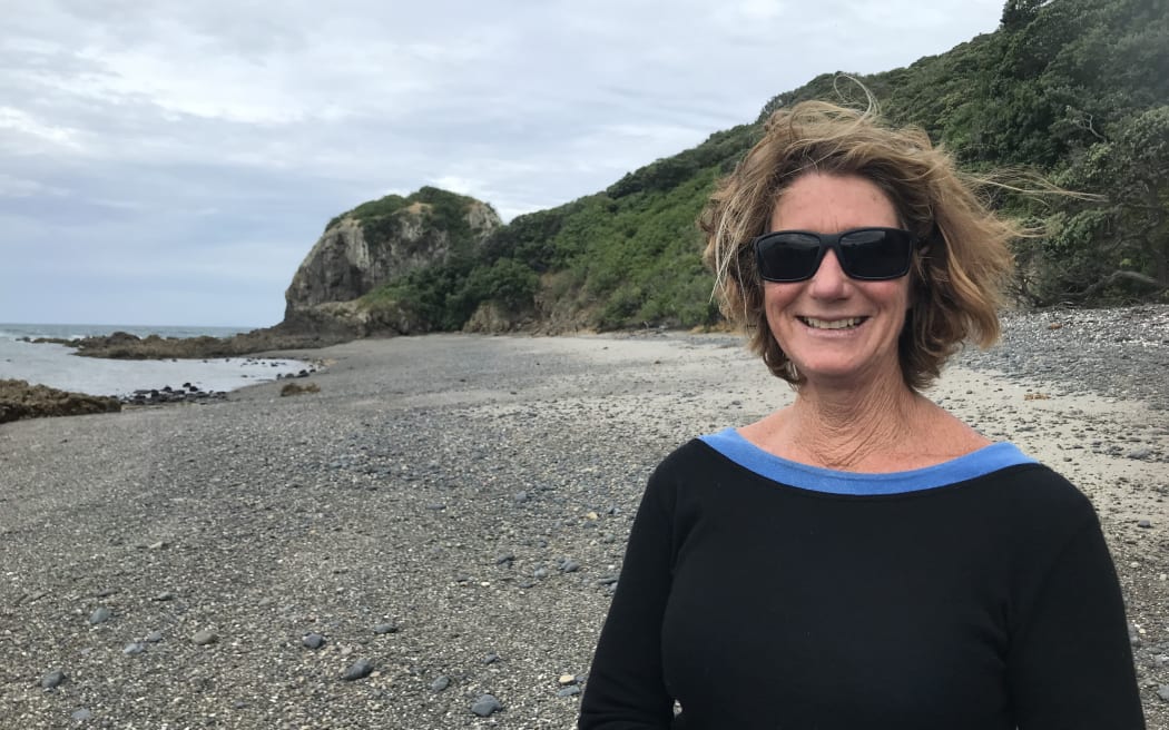 Sue is standing on a gravelly beach. She is smiling and wearing sunglasses.