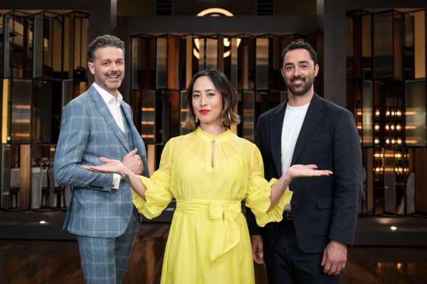 Melissa Leong, in a bright yellow dress, holds out her hands to present two smiling bearded men in suit jackets.