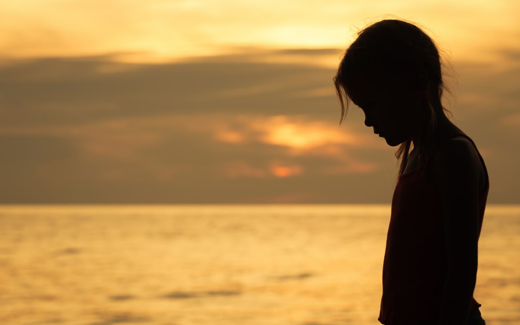 Portrait of sad blond little girl standing on the beach at sunset time.