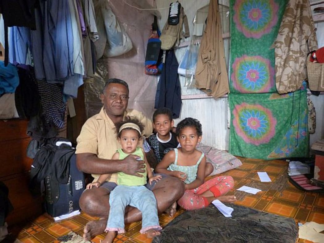 Tevita and young grandchildren in a room with a couple of mattresses on floor and clothes hanging on walls in their home in a squatter settlement in Suva