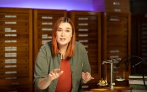 A still from a video. Morgane is gesturing to the camera while she talks. She stands behind a desk in an office with bugs on displays, in boxes and on scales.