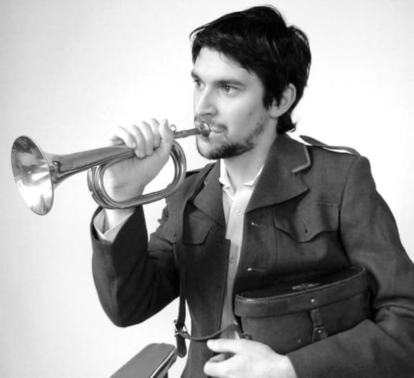 Dudley Benson wears a military jacket and plays a trumpet while looking to the side.