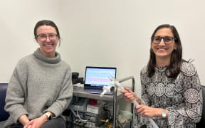 Two women with long dark hair and glasses smile and sit next to a laptop on a metal trolley in front of a white wall. On the tray underneath the laptop is a complex machine with a long tube, that the woman on the right is holding in her hands.