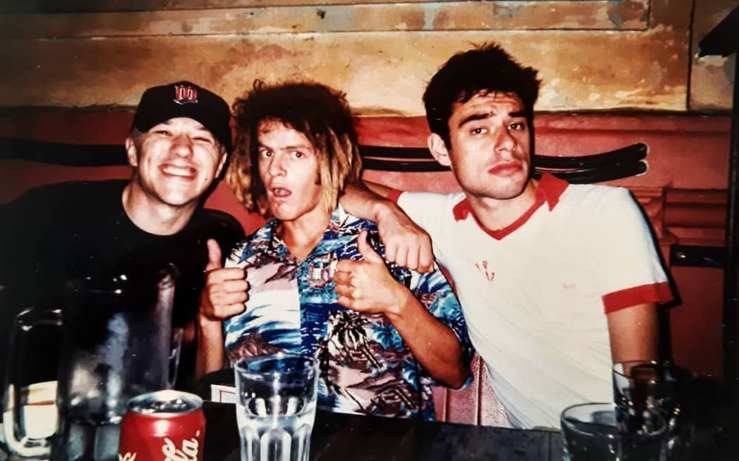A young DJ StickyFingaz rocking a Hawaiian shirt, and some brown dreadlocks with bleached ends. He's accompanied by two other younger men.