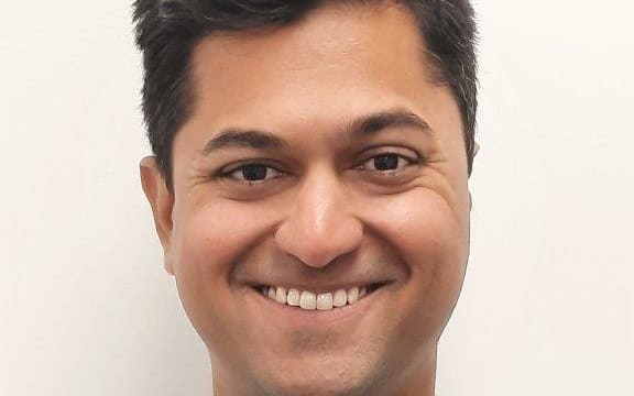 A headshot of a man wearing a white and blue checkered shirt. He is smiling directly at the camera.