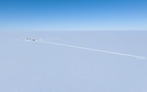 A drone image of the white expanse of flat ice beneath a clear blue sky with a tiny camp off to the left, and a long trail in the ice extending to the right.
