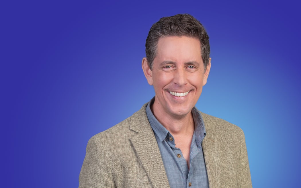 Host Wallace Chapman in a blue shirt and casual jacket. Soft blue background.