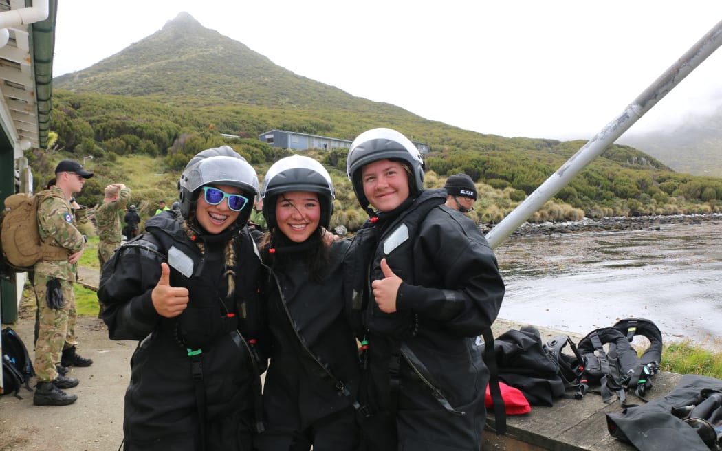 Three young women are wearing black dry suits, lifejackets and helmets. They are smiling and two of them are showing thumbs up. They are on a wharf and behind them rises Beeman hill and you can see army personnel and the MetService hostel.