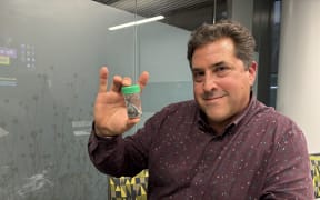 A man in a purple shirt with a grey pattern holds up a small plastic vial with a green lid. Inside the vial are around six silvery fish specimens. The man is looking at the camera and smiling.