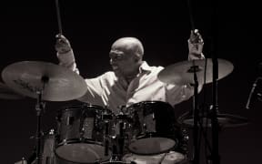 A black and white photo of drummer Roy Haynes in action, lifting his drumsticks in the air and sitting behind a drum set.