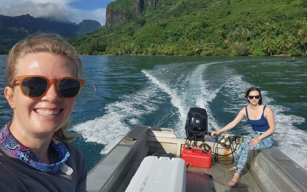 A selfie taken by a woman wearing sunglasses on a boat, with another woman visible on the right sitting next to and operating the boat's motor. Behind them, the wake of the boat extends across blue ocean and steep green hillsides covered with vibrant green tropical forest. The sky is cloudy but low clouds cling to the tops of the hills and rock formations.