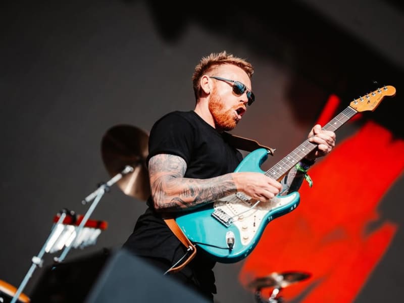Joel Shadbolt, a red-headed and bearded man in a black t-shirt, raises his teal blue electric guitar to the sky.