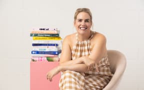 An image of Christchurch-based food writer and chef Sam Parish. Sam is wearing a brown and white checked jumpsuit and sitting next to a stack of cookbooks.