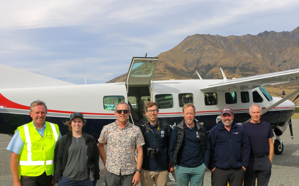 Seven people stand in a line smiling at the camera in front of a small plane with a mountain in the background.