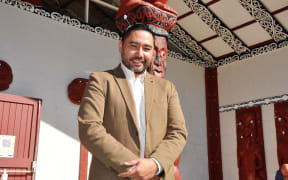 Te Matapihi General Manager, Wayne Knox, stands in front of a wharenui wearing a white collared button down shirt and a tan blazer.