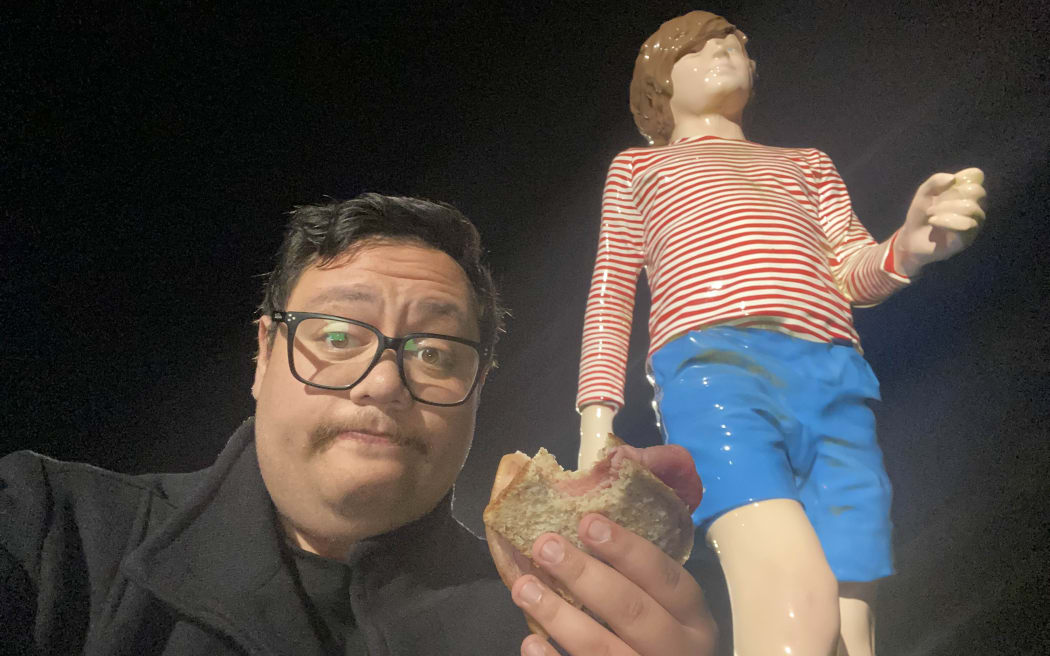 A man in a coat holds up a freshly cooked bacon sandwich up to a giant statue of a kid in blue shorts and a red and white striped shirt.