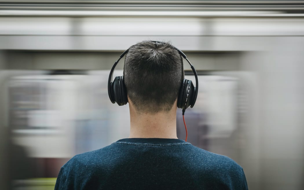 A man wearing headphones facing away from the camera, with a moving train blurred in the background.