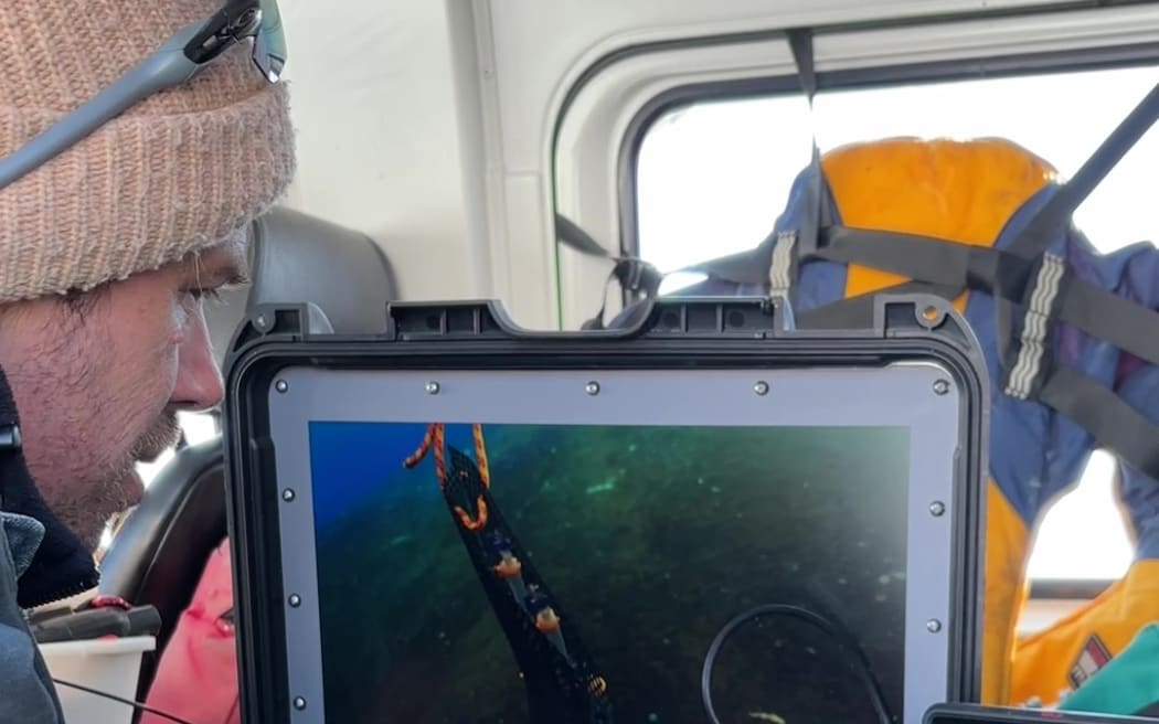 A man wearing a beanie and jacket sits at a monitor showing a view of the seafloor. He appears to be sitting in a vehicle of some kind.