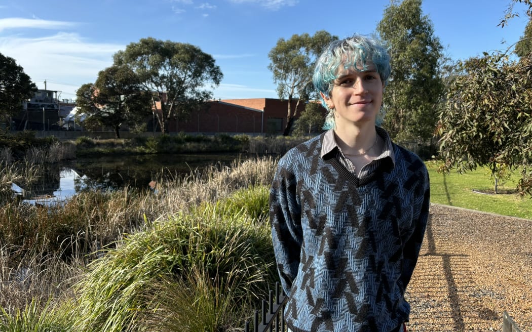 A man with blue hair wearing a blue and black sweater stands in front of vegetation beneath a blue sky. A large building is visible in the background. The man is smiling at the camera and has his hands behind his back.