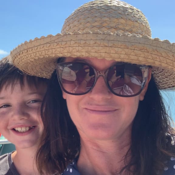 A portrait of Emily in a sun hat and glasses with a boy over her right shoulder, with a backdrop of bright blue sky. She is smiling and wearing a blue polka dot top.
