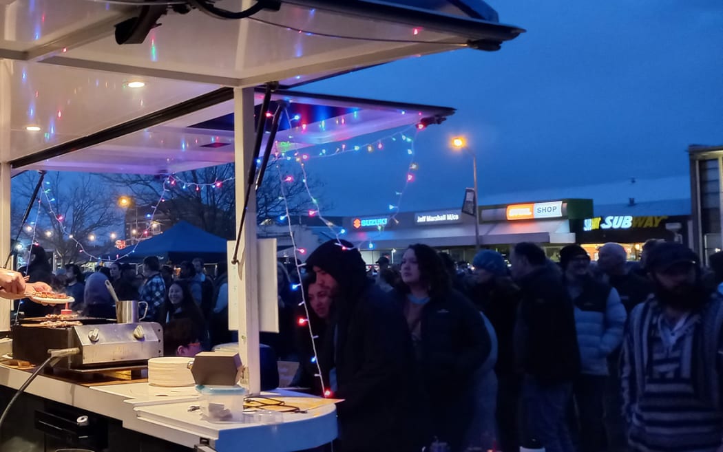 A bustling market at twilight. A food truck strung with fairy lights is busy making orders.