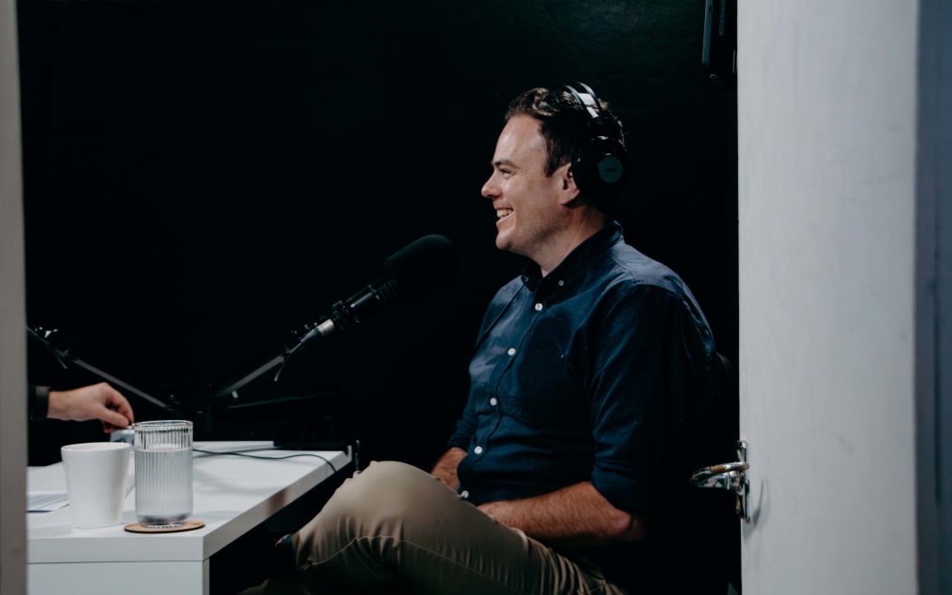 An image of Lloyd Burr sitting at a white desk speaking into a microphone. The image is taken through a doorway.