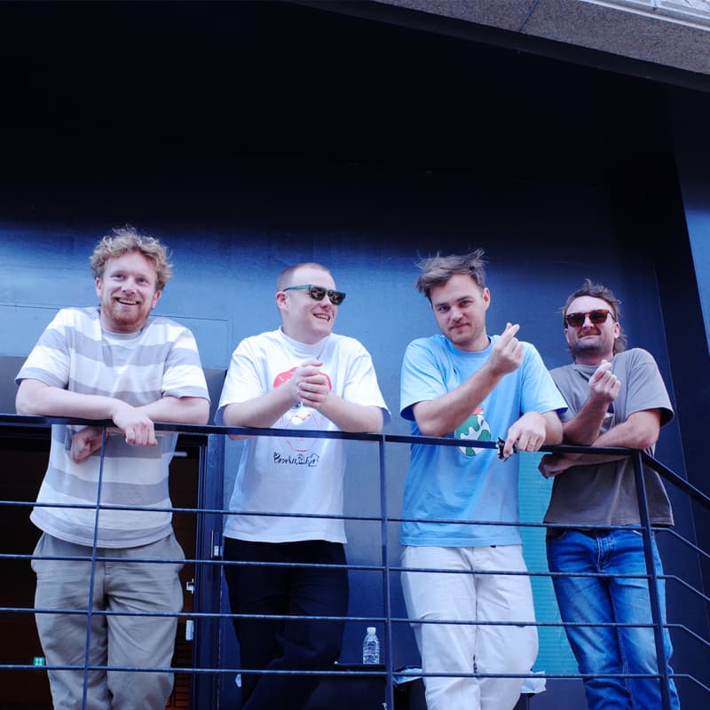 Four men wearing t-shirts stand in a row on a balcony.