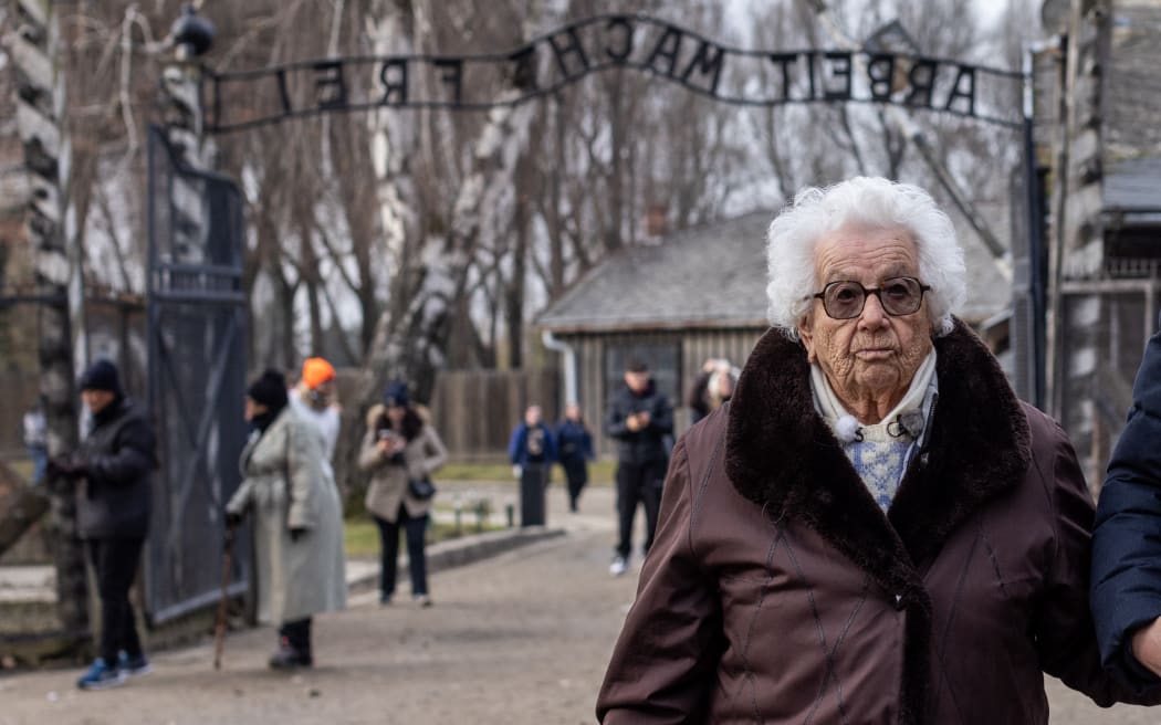 Holocaust survivor Esther Senot, 97, helped by unidentified man enters through the guide in front of the gate with "Arbeit macht frei" (Work sets you free) written across at the entrance to the Auschwitz-Birkenau former German Nazi concentration and extermination camp in Oswiecim, southern Poland, on 5 December 2024. - She was 15 years old when she arrived to the Nazi Auschwitz-Birkenau camp in September 1943 where she has now returned to tell her story to a group of nearly a hundred second-level school students from France. (Photo by Wojtek RADWANSKI / AFP)
