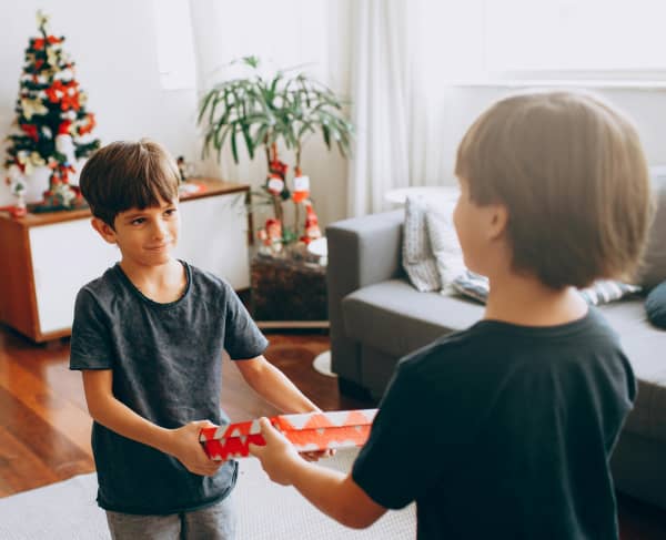 A young boy in a black t-shirt hands a Christmas present to a taller boy who is also in a black t-shirt.
