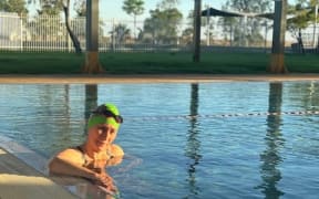 Janene is in an outdoor lap pool with a swimming cap and goggles on.