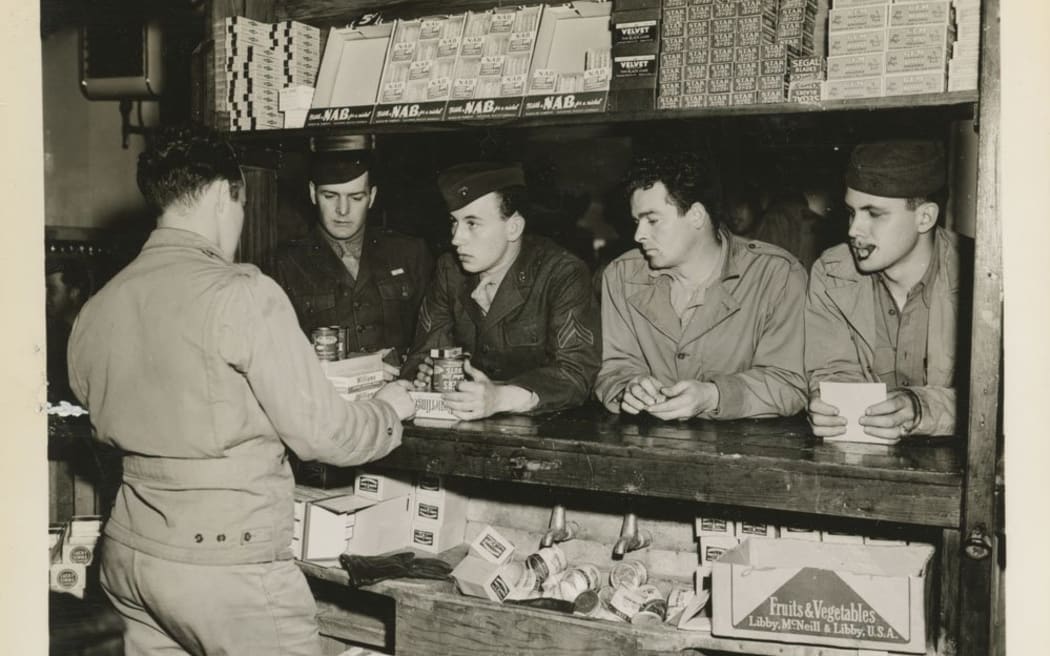 An old photograph. Four US Marines sit at a bar bench, being served by a clerk. They all wear fatigues and hats.