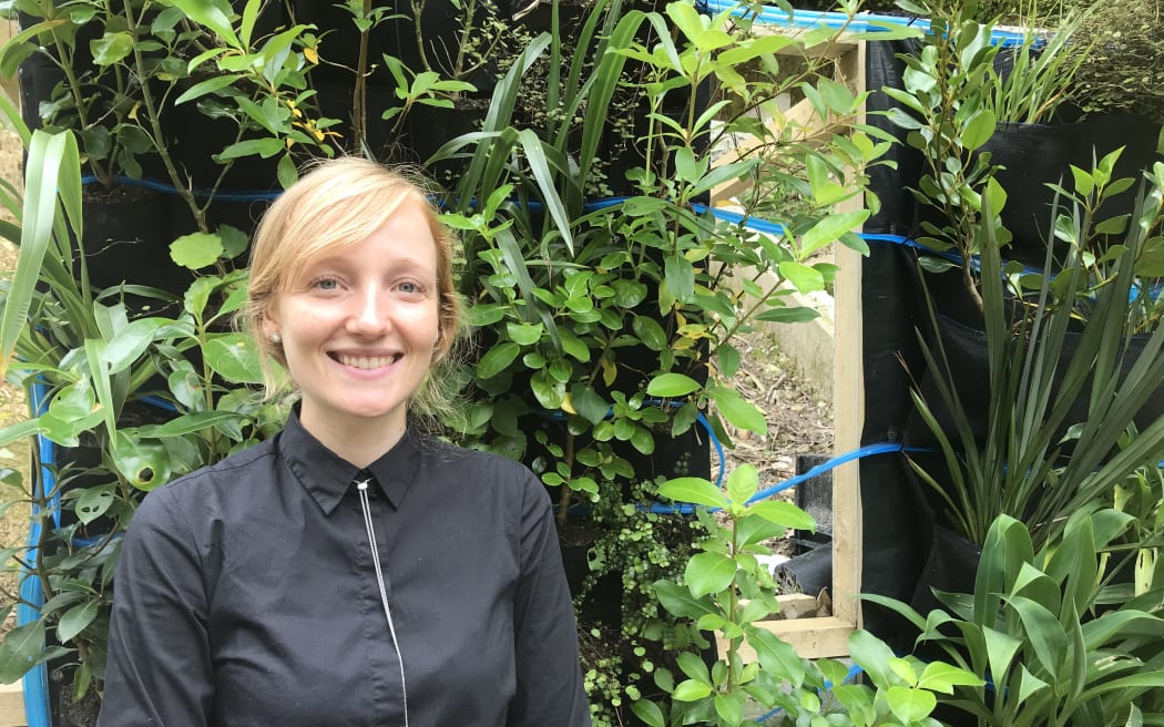 Maggie McKinnon is standing in front of a vertical garden of lush green plants. She is smiling.