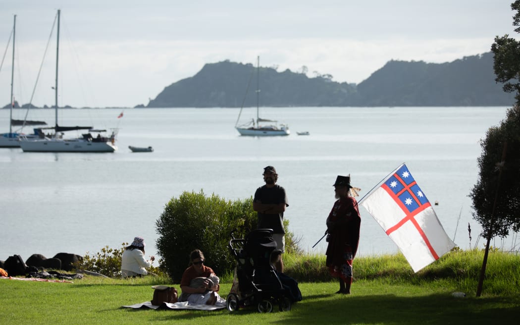 Crowds gather in the grassy areas around the Waitangi Forum Tent including one person who is holding a United Trible flag on a pole. Te Ti Bay is pictured in the background.