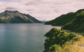 A road runs through scrub alongside a lake with mountains in the background and a cloudy sky.