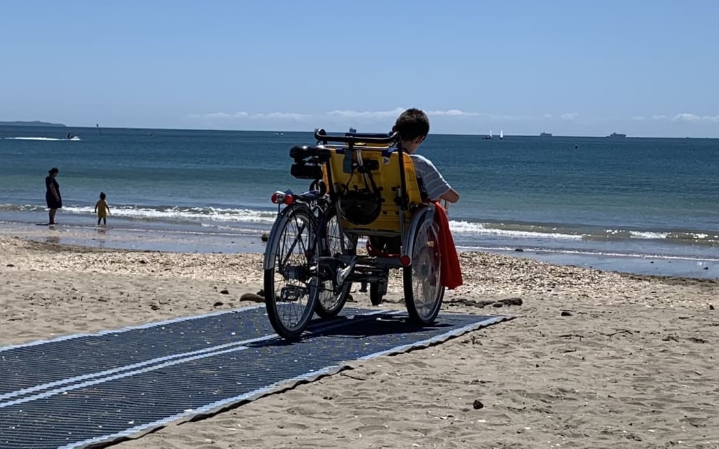 Finlay Graham on a wheelchair bicycle on a beach mat at Takapuna Beach.
