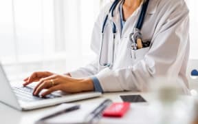 A woman in a white coat and stethoscope on her shoulders types on a laptop.