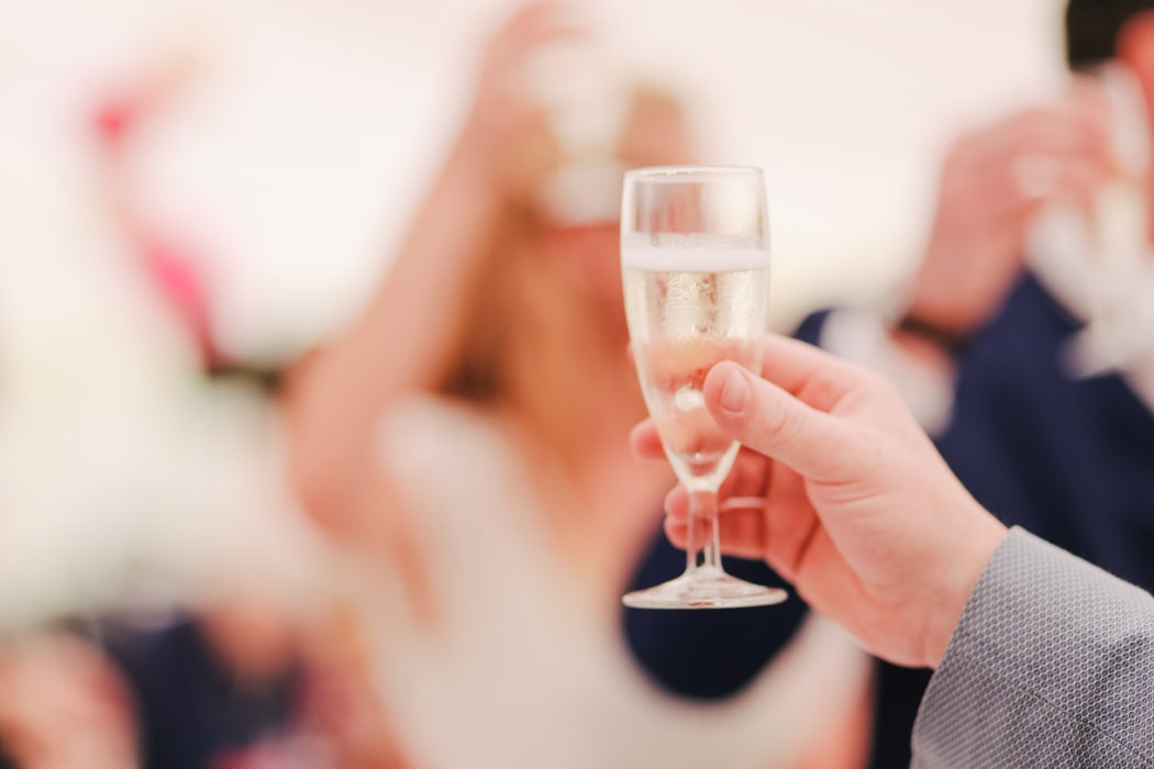 Glass of champagne being held up in celebration iwth partygoers in background.