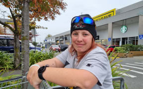 Olivia Bovey stands outside a supermarket leaning against a trolley. She is wearing a grey t-shirt, black beanie and has sunglasses on top of her head.