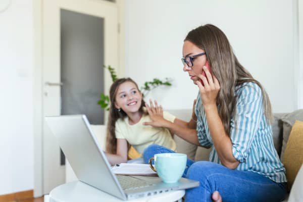 A woman speaking on the phone and on her laptop holds out her hand to stop a young girl next to her from interrupting.