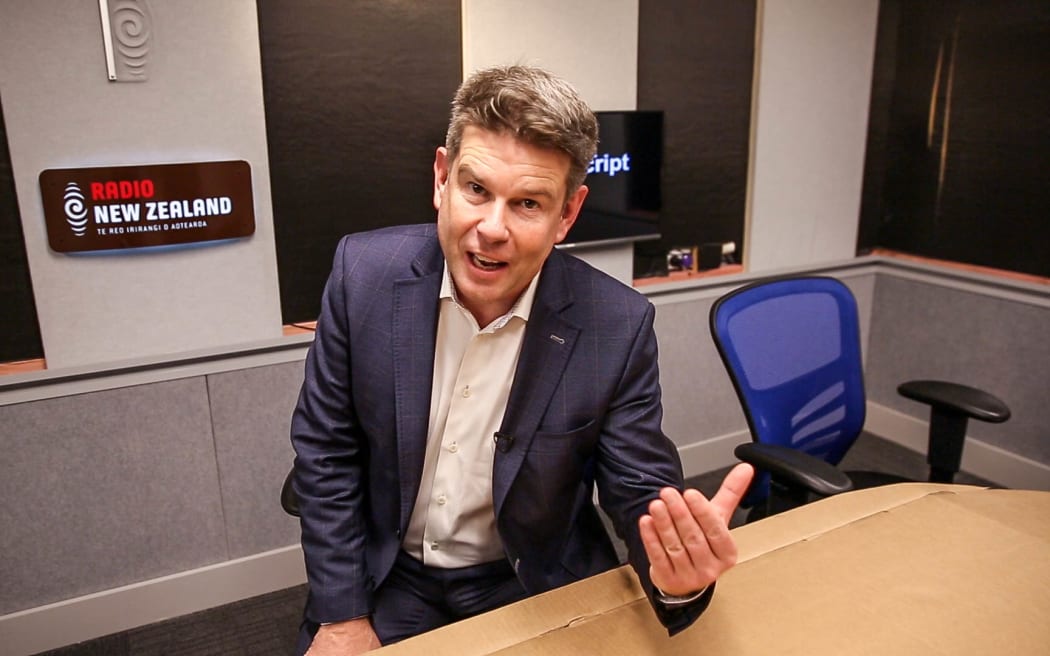 John Campbell sitting at a temporary cardboard desk in the Checkpoint studio at Radio New Zealand.