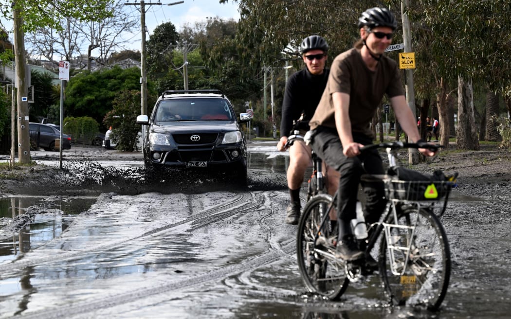People cycle and cars drive down mud-strewn streets affected by floods in the Melbourne suburb of Maribyrnong on October 15, 2022. - Australia reported the first fatality from days of widespread flash flooding on October 15, 2022 despite heavy rains easing and flood levels topping out across much of the southeast. Hundreds of homeowners began a long clean-up after storm waters engulfed streets, houses and cars across three states, with Melbourne suburbs among the worst hit. (Photo by William WEST / AFP)