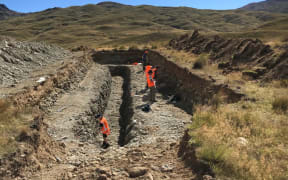 The trench as viewed from the top, with the team at work. Ashleigh is cleaning the lower trench area. Mark, James, Govinda and Andy are looking at the gridded/marked right hand side of the trench.