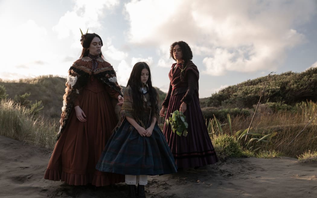 MĀRAMA (Ariāna Osborne), ANAHERA (Evelyn Towersey) and PEGGY (Umi Myers) stand infront of the granite headstone.