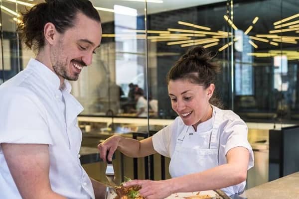 Two smiling people in white shirts work in a kitchen together.
