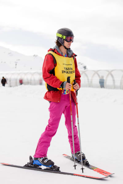 Presenter Ari Kerssens is dressed in a yellow bib that says 'vision impaired'. He is holding skis while stood on a snowy slope.